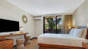 Une chambre d’hôtel avec un lit, un bureau avec une télévision, un miroir et une vue sur les palmiers par la fenêtre.
