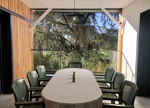 Uma sala de jantar moderna com uma mesa grande, seis cadeiras e vista para árvores através de amplas janelas.