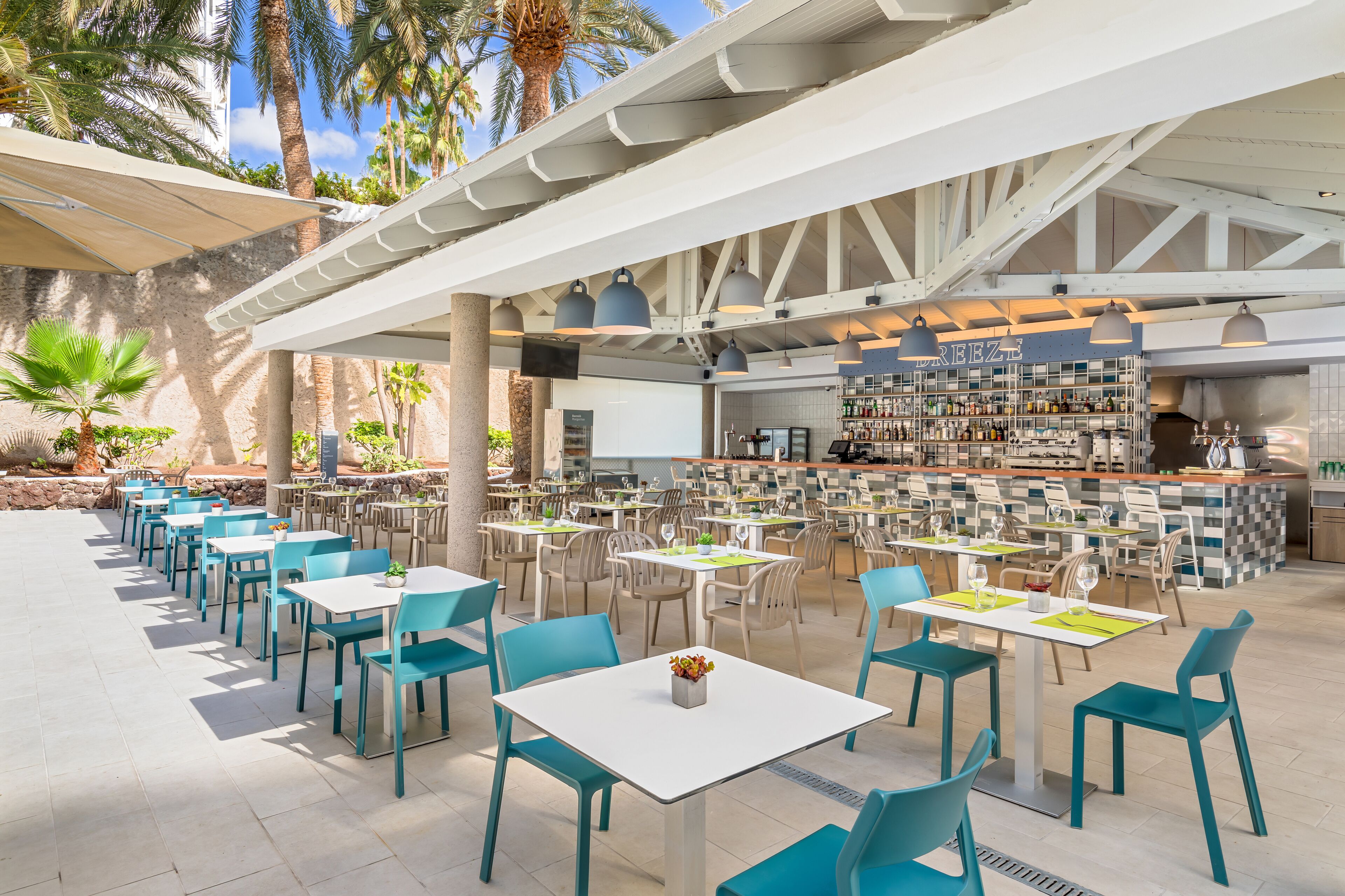 An outdoor dining area with multiple tables and chairs, a bar area with shelves of bottles, and palm trees in the background.