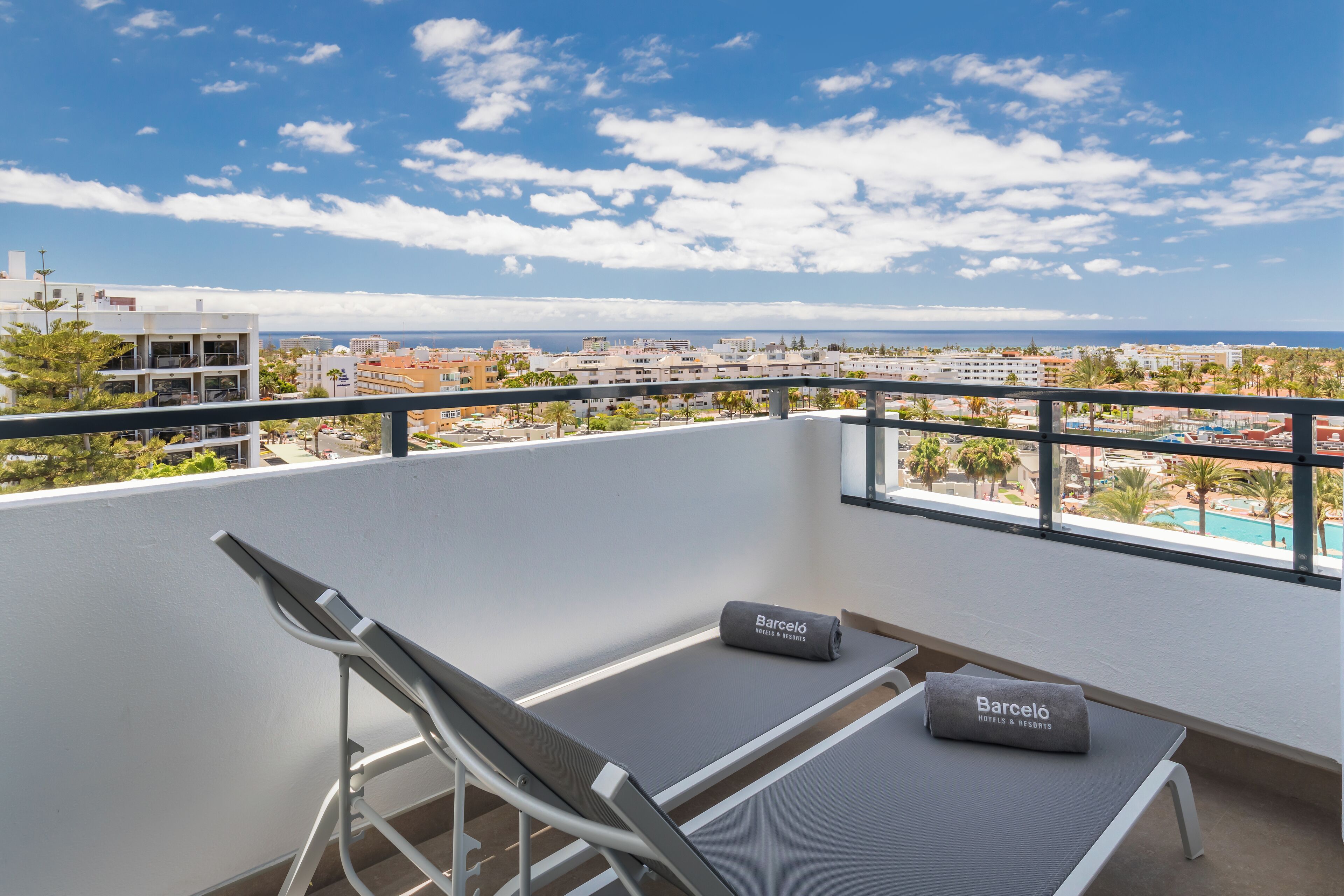 Un balcón con vista a la ciudad y al mar, una silla, una mesita y un cojín con el nombre del hotel.