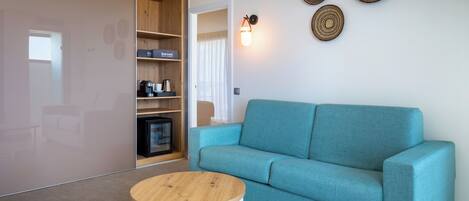A modern living room with a teal sofa, a wooden coffee table, and a built-in shelving unit with a coffee maker and a small TV.