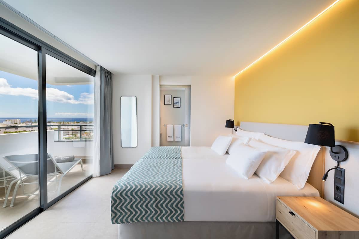 A modern hotel room with a large bed, bedside tables, a nightstand, a balcony with a glass door, and a view of the cityscape.