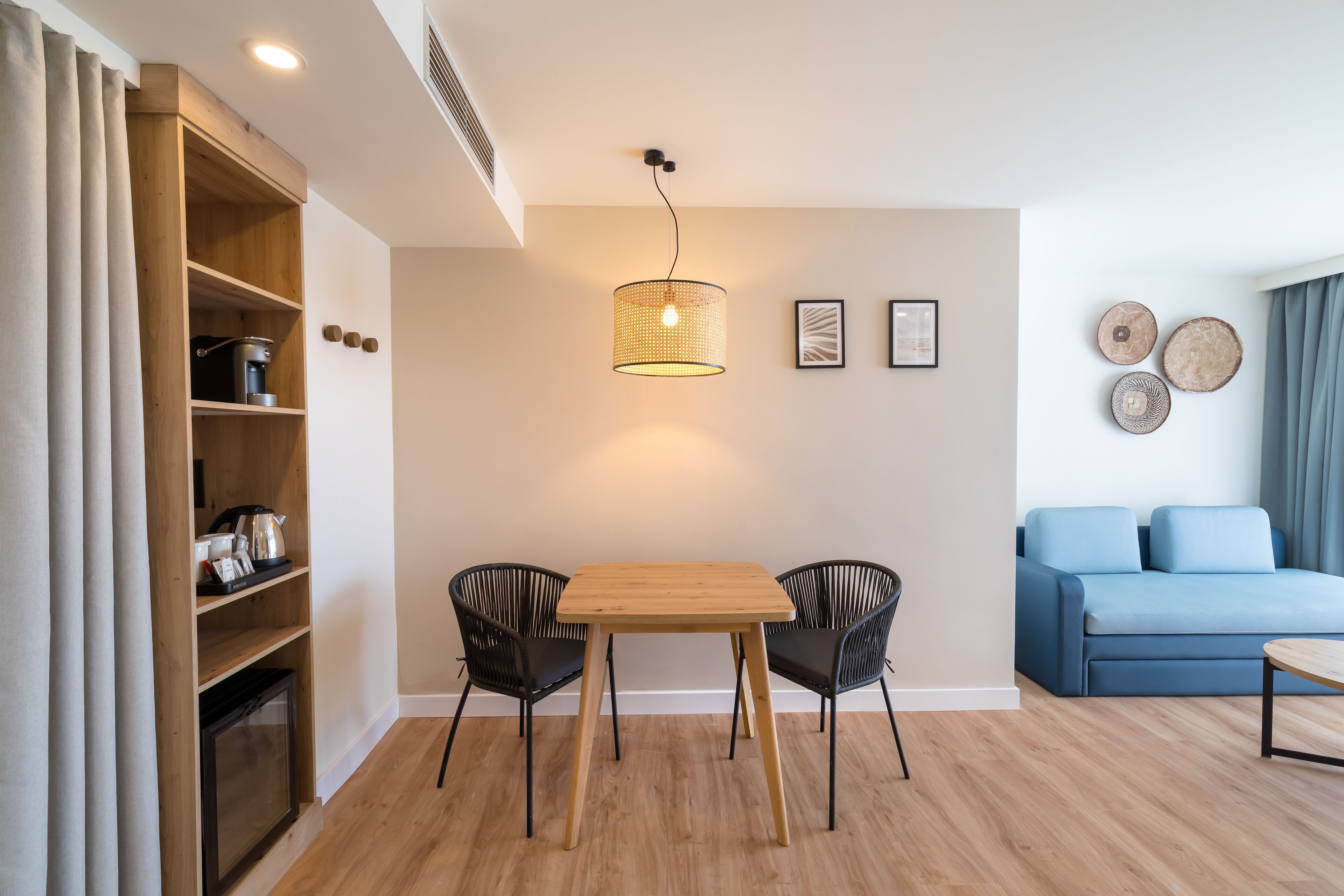 Una sala de estar moderna con una mesa de comedor de madera, dos sillas negras, un sofá azul y una máquina de café sobre una estantería de madera.
