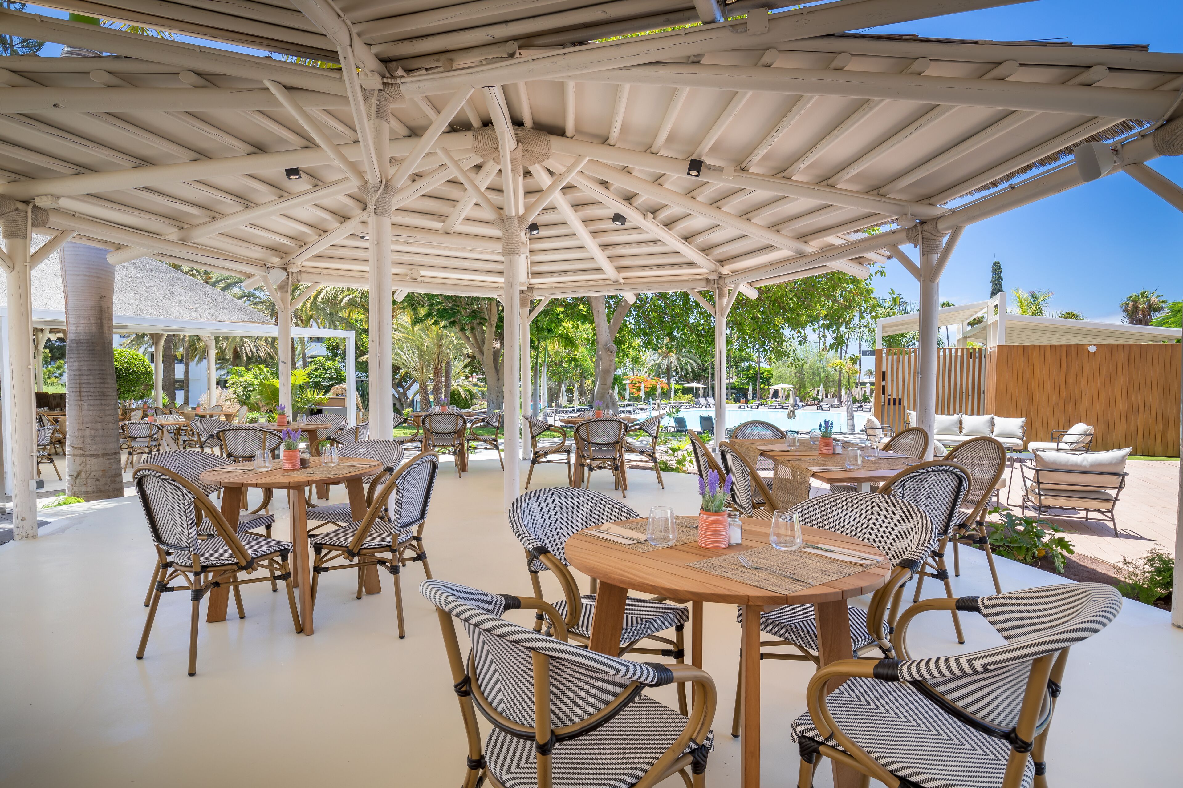 Une terrasse extérieure avec des tables en bois et des chaises en osier, entourée d’une zone couverte dotée d’un toit blanc.