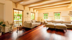 A spacious living room with wooden flooring, a beige sofa, a round coffee table, and large windows offering a view of greenery outside.