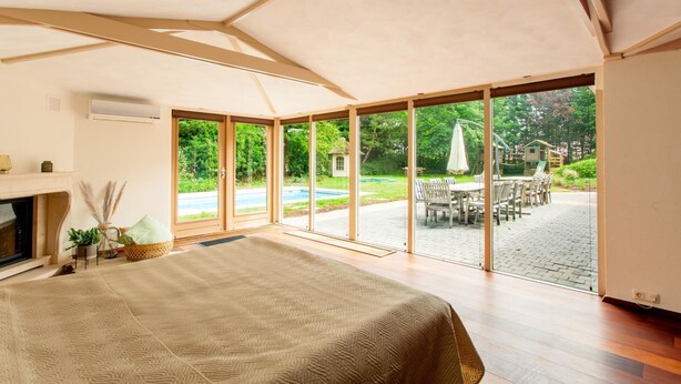 A spacious bedroom with a large bed, a fireplace, and a view of a backyard with outdoor furniture and a swimming pool.