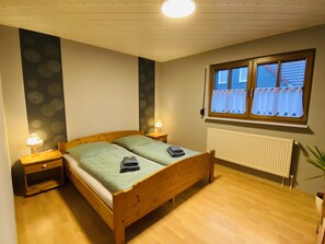 A bedroom with a wooden bed, two bedside tables with lamps, a window with curtains, and a radiator.