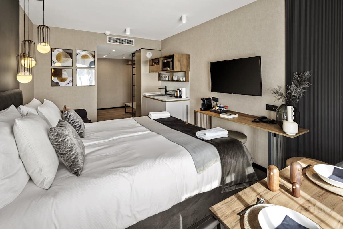 A modern hotel room with a large bed, a desk with a flat-screen TV, a kitchenette, and a dining area.