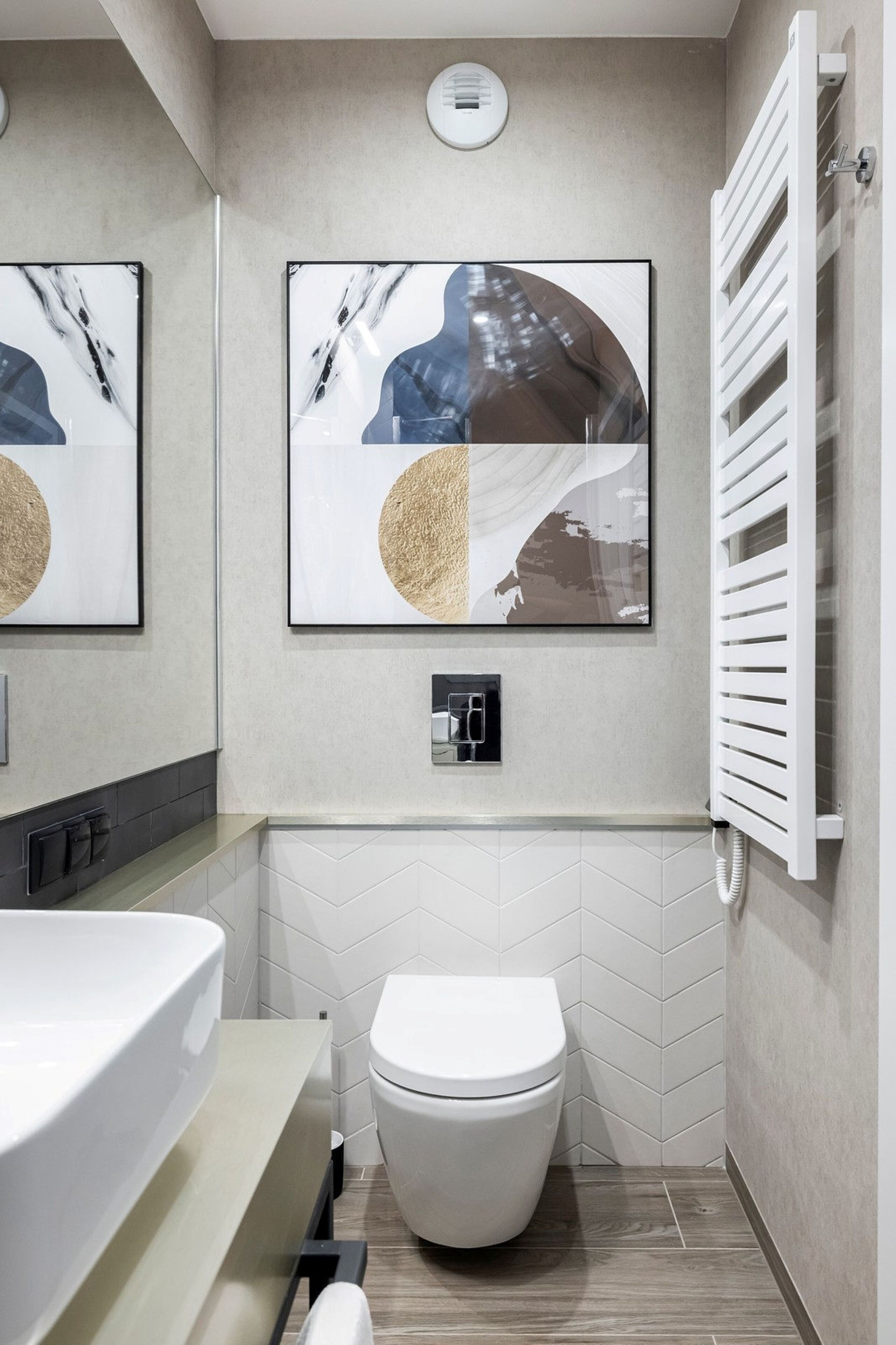 Een moderne badkamer met een wit toilet, een wastafel en een handdoekhouder. Twee kunstwerken in lijsten hangen aan de muur en er is een witte radiator.