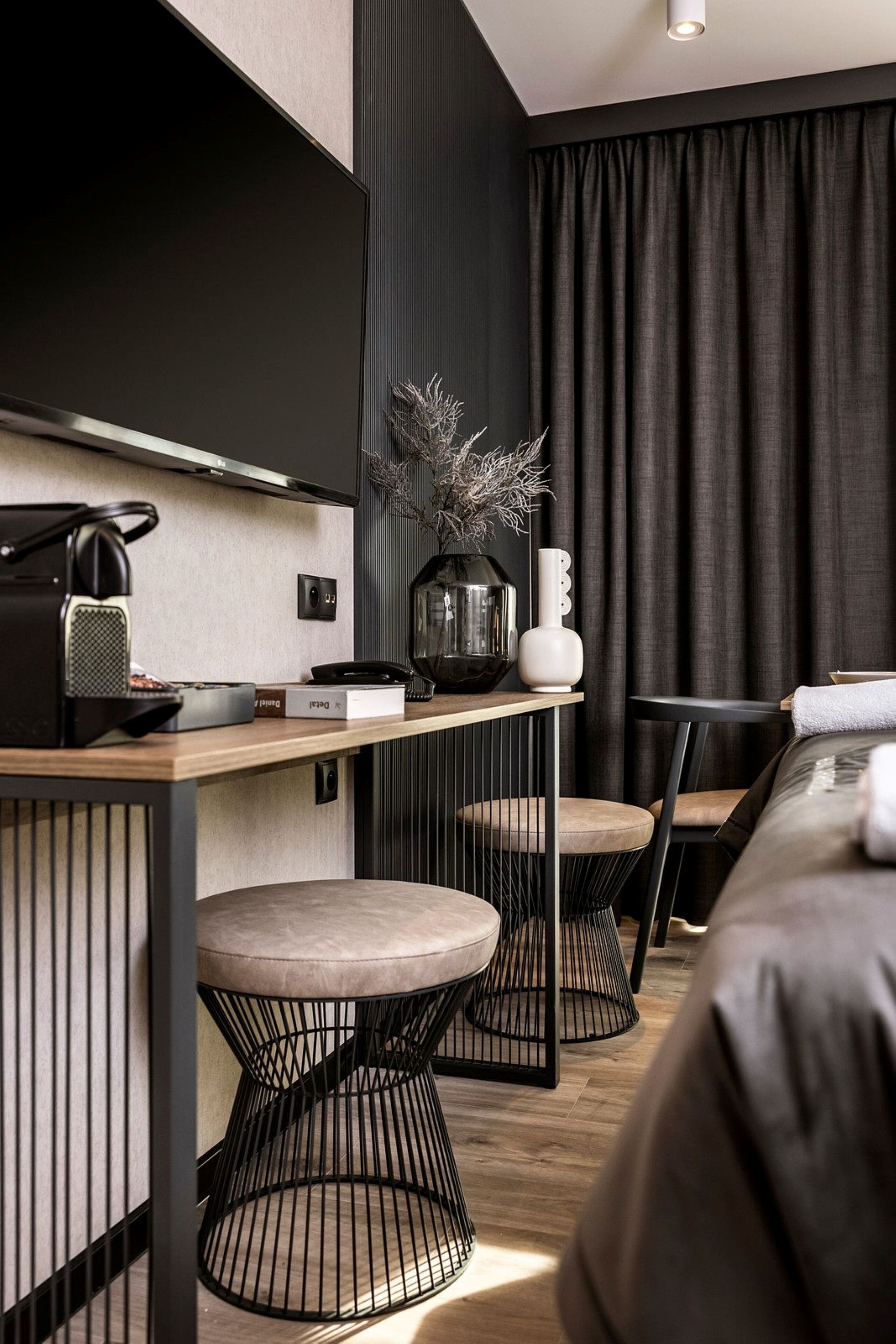 A modern hotel room with a flat-screen TV mounted on the wall, a wooden desk with a telephone, a vase with dried plants, and a bed with a grey headboard.