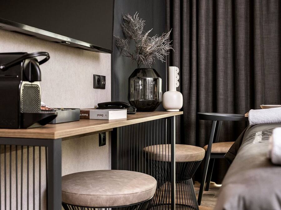 A modern hotel room with a flat-screen TV mounted on the wall, a wooden desk with a telephone, a vase with dried plants, and a bed with a grey headboard.