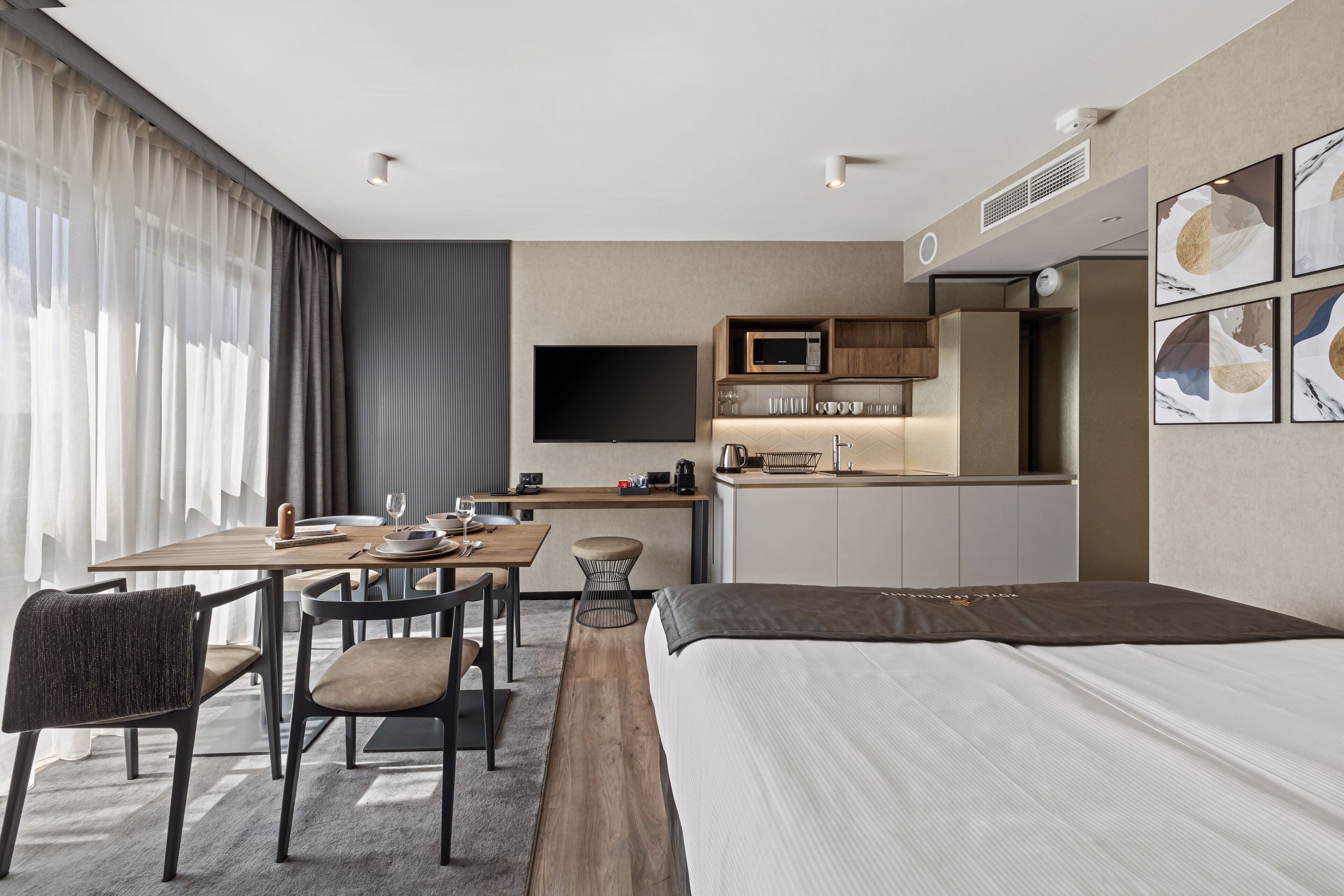 A modern hotel room with a large bed, a dining area with a table and chairs, a kitchenette, and a flat-screen TV mounted on the wall.