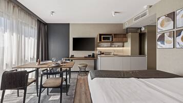 Una habitación de hotel moderna con una cama grande, una zona de comedor con mesa y sillas, una kitchenette y un televisor de pantalla plana montado en la pared.