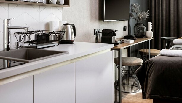 Une chambre d’hôtel moderne dotée d’un coin cuisine, d’un téléviseur à écran plat fixé au mur et d’un lit avec une tête de lit de couleur foncée.