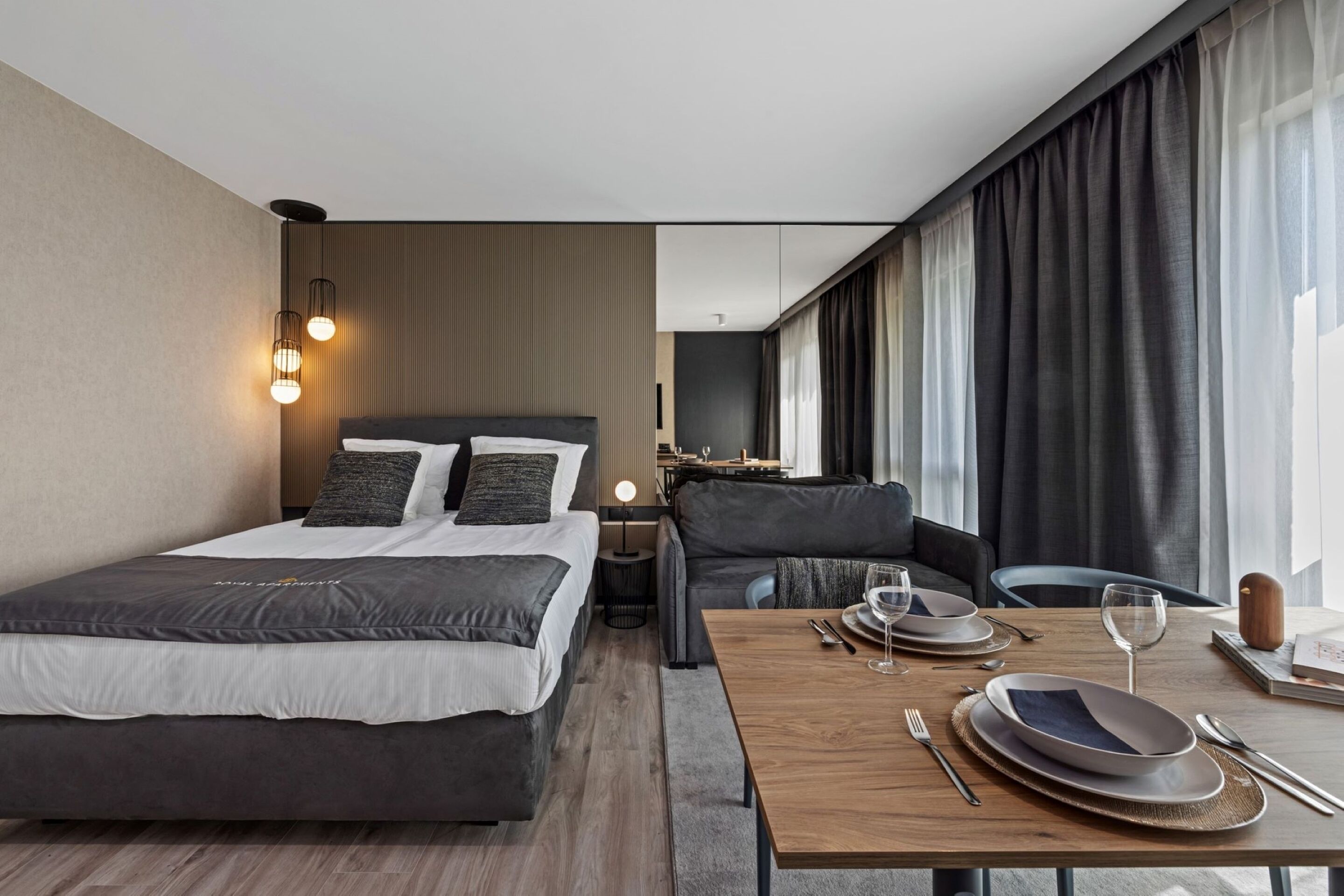 Una habitación de hotel moderna con cama, sofá, mesa de comedor para dos personas y un gran ventanal con cortinas.