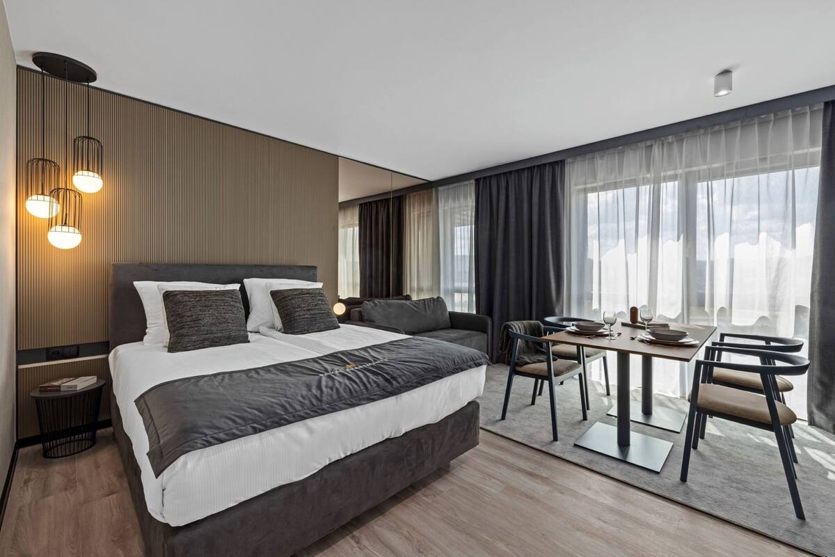 A modern hotel room with a large bed, a dining area with a table and chairs, and large windows with curtains.