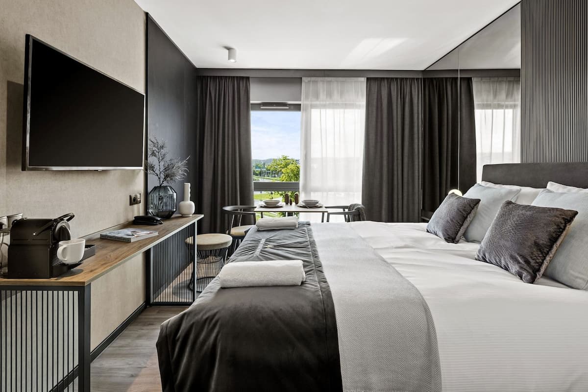 A modern hotel room with a large bed, a desk with a coffee maker, a TV, and a view of the outdoors.