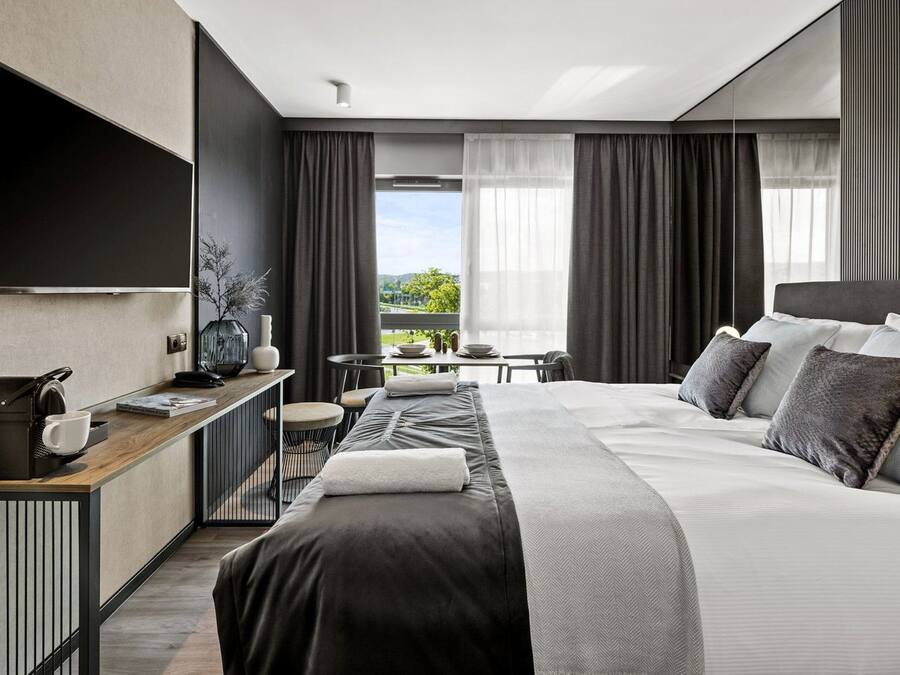 A modern hotel room with a large bed, a desk with a coffee maker, a TV, and a view of the outdoors.