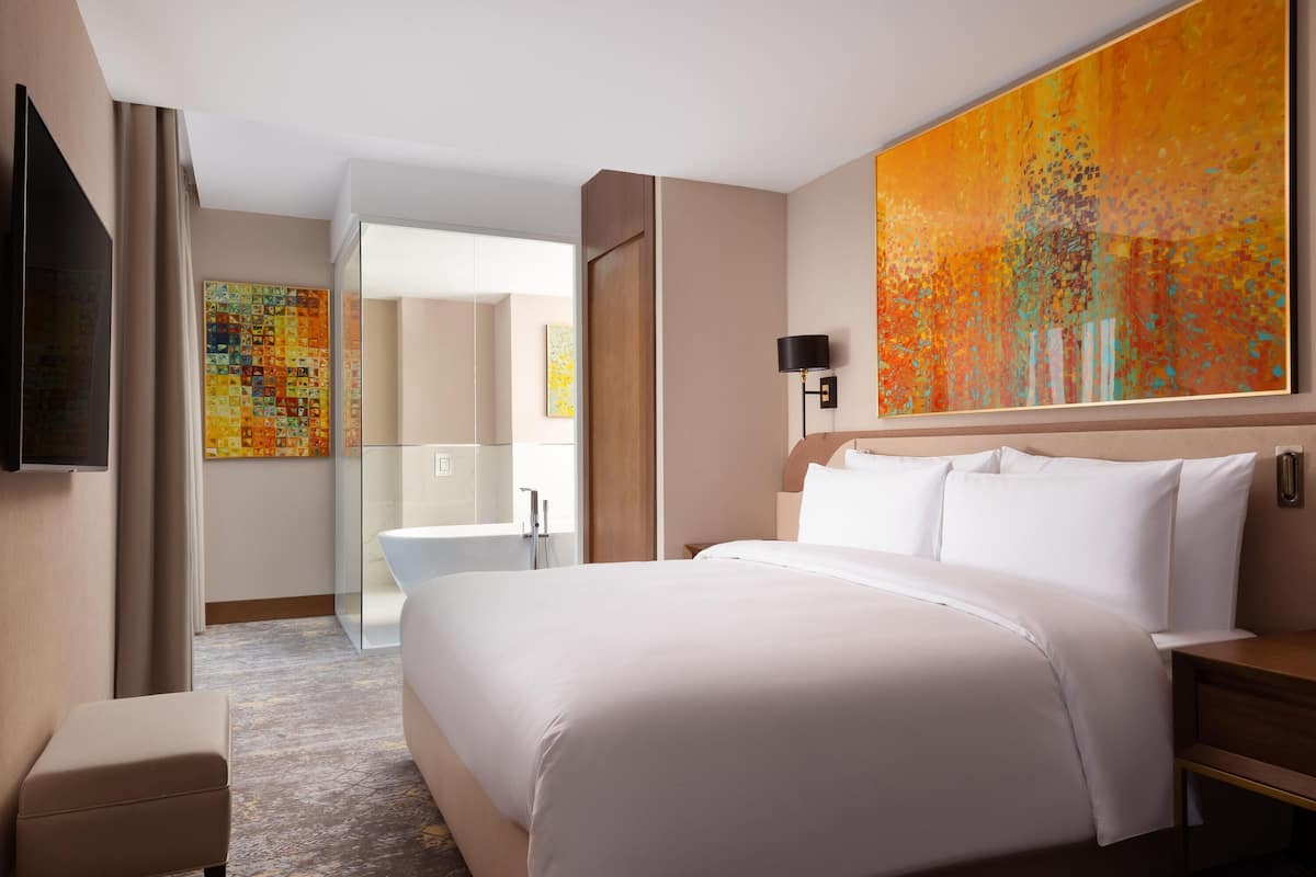 Une chambre d’hôtel avec un grand lit, une table de chevet, une télévision et une salle de bain avec baignoire, ainsi qu’un grand tableau abstrait accroché au mur.