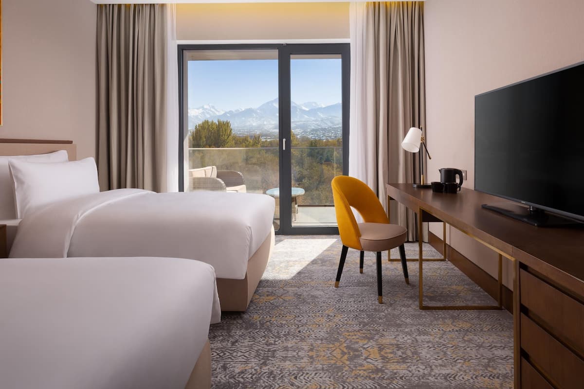 Une chambre d’hôtel avec deux lits, un bureau avec une télévision et un balcon offrant une vue sur la montagne.