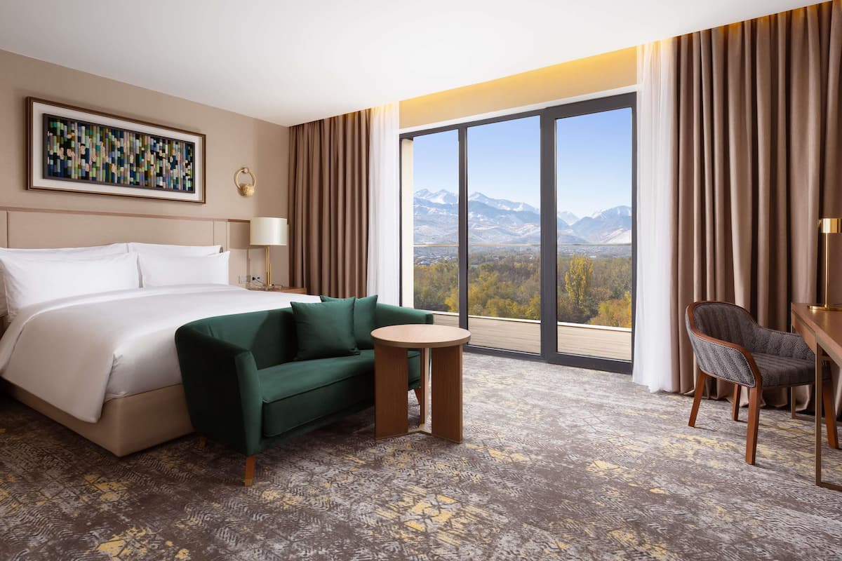 Une chambre d’hôtel avec un lit, un canapé vert, une petite table ronde, une chaise et une grande fenêtre donnant sur la montagne.
