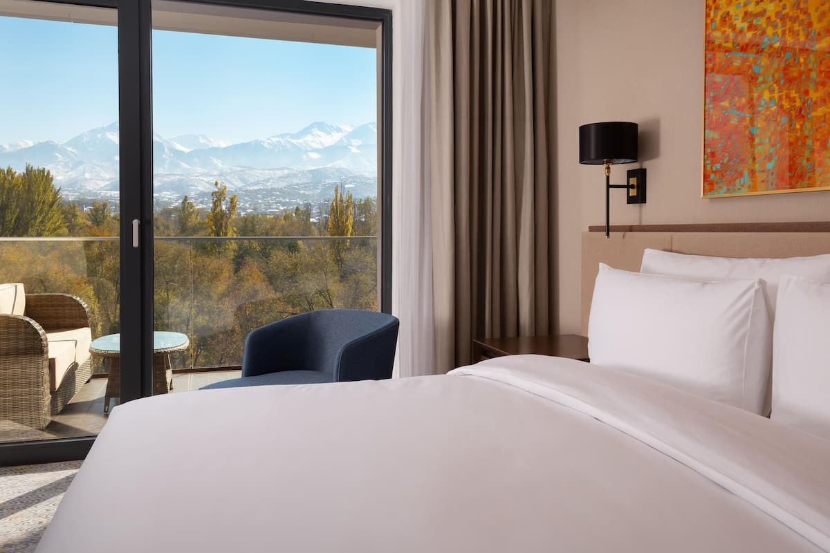 Une chambre d’hôtel avec un grand lit, une chaise, une table, une vue sur les montagnes et un tableau au mur.