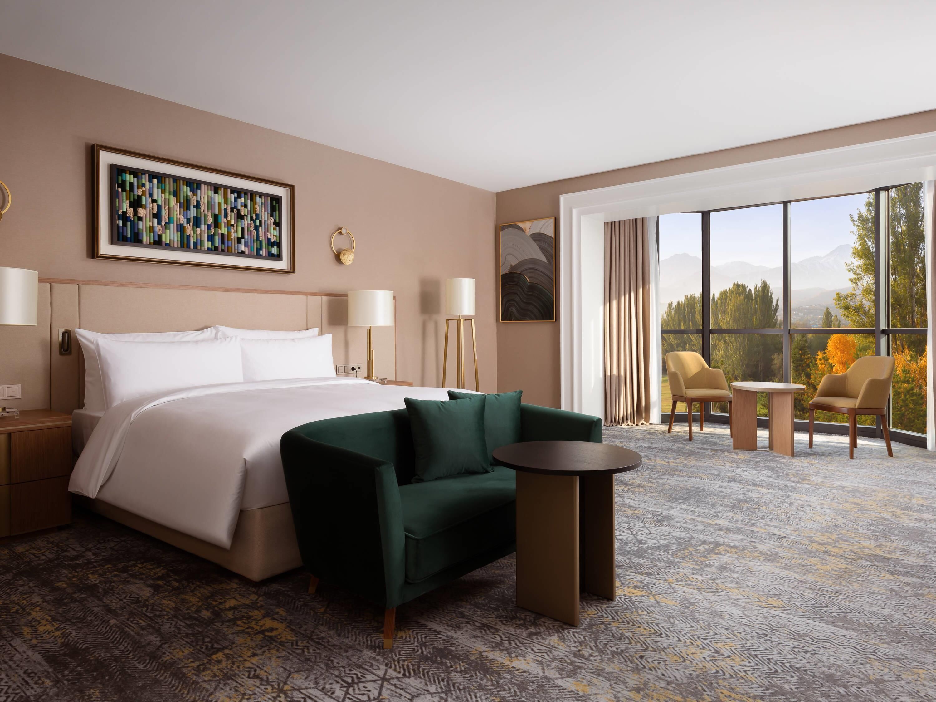 Ein Hotelzimmer mit einem Bett, einem grünen Sofa, einem kleinen Tisch, einem großen Fenster mit Blick und einem Gemälde an der Wand.