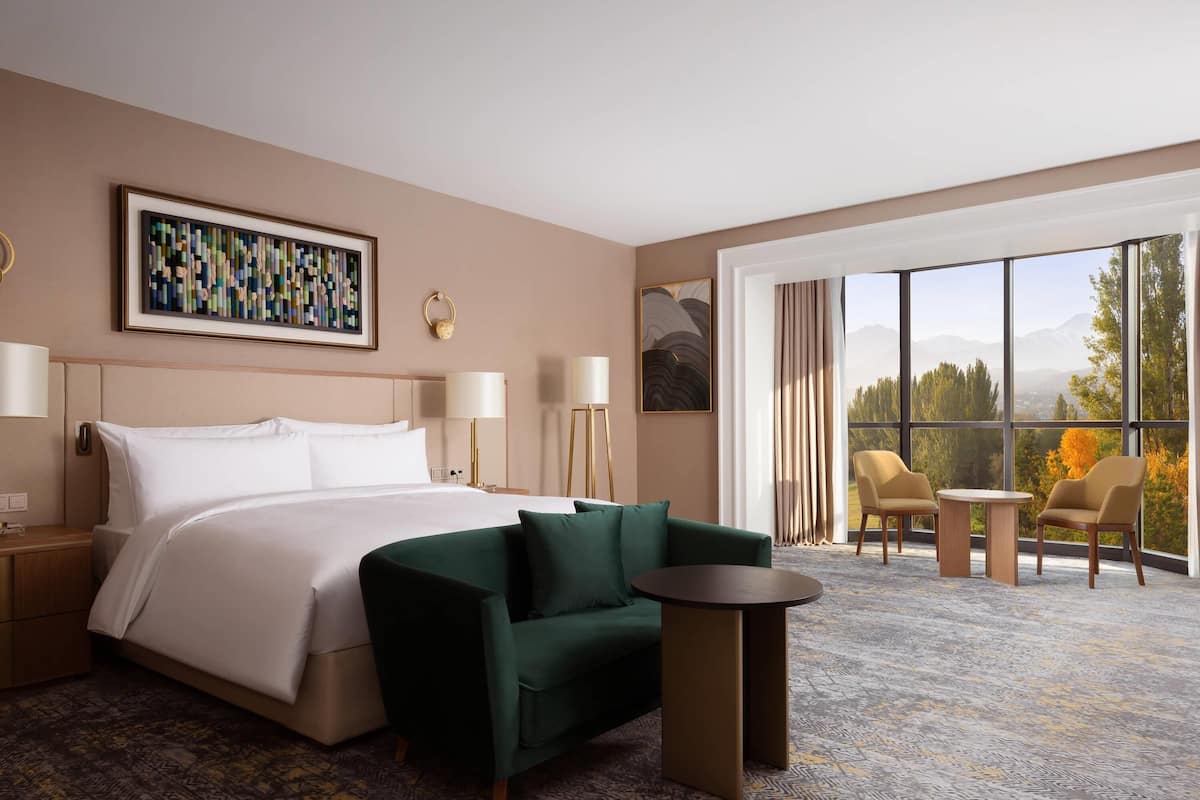 Une chambre d’hôtel avec un lit, un canapé vert, une petite table, une grande fenêtre donnant sur un beau paysage et un tableau accroché au mur.
