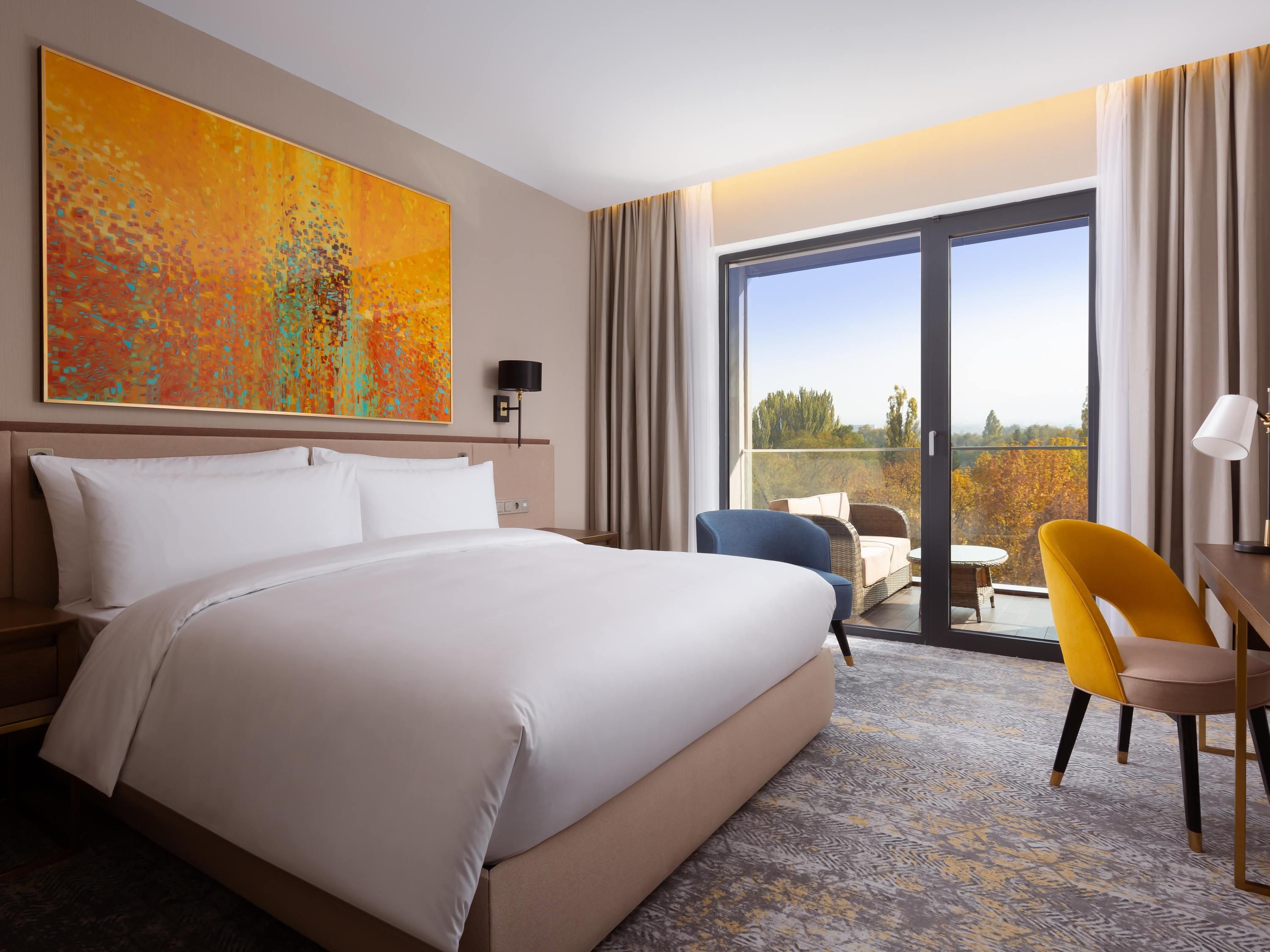 A hotel room with a large bed, a desk, a chair, a window with a view, and a painting on the wall.