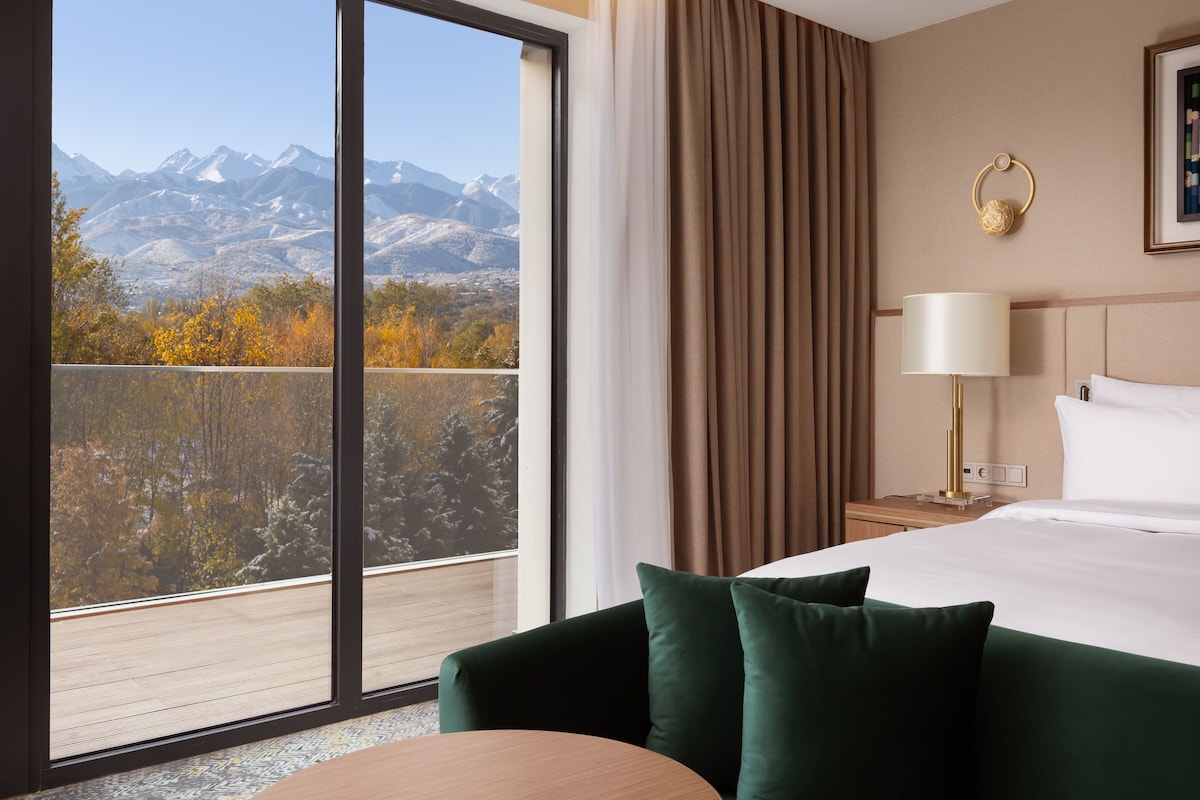 Une chambre d’hôtel avec une grande fenêtre, un lit, une table de chevet, une lampe et une vue sur les montagnes.