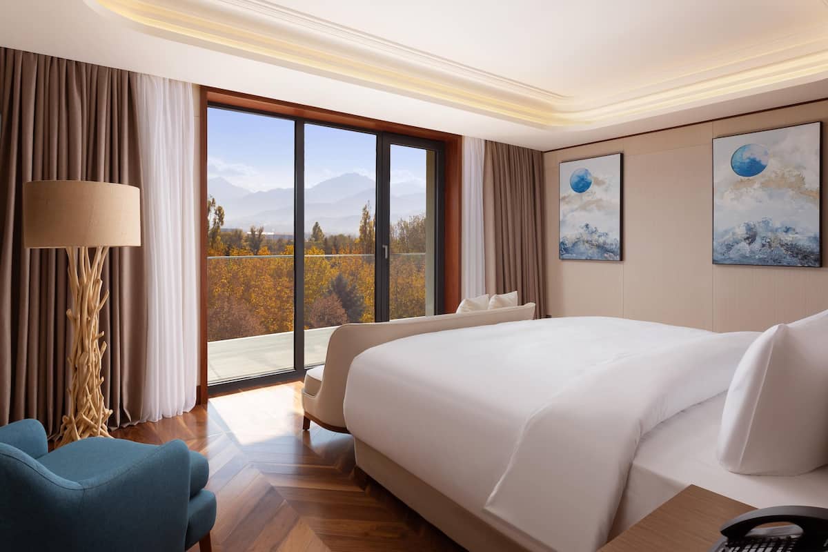 Une chambre d’hôtel avec un grand lit, un bureau avec un téléphone, une chaise, une lampe et une vue sur les arbres et les montagnes par la fenêtre.