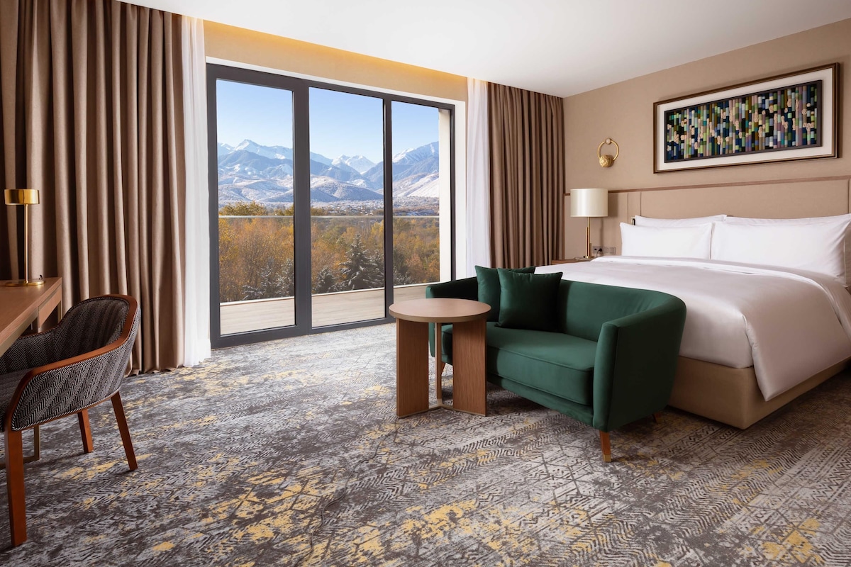 A hotel room with a large bed, a green armchair, a small table, and a view of mountains through floor-to-ceiling windows.