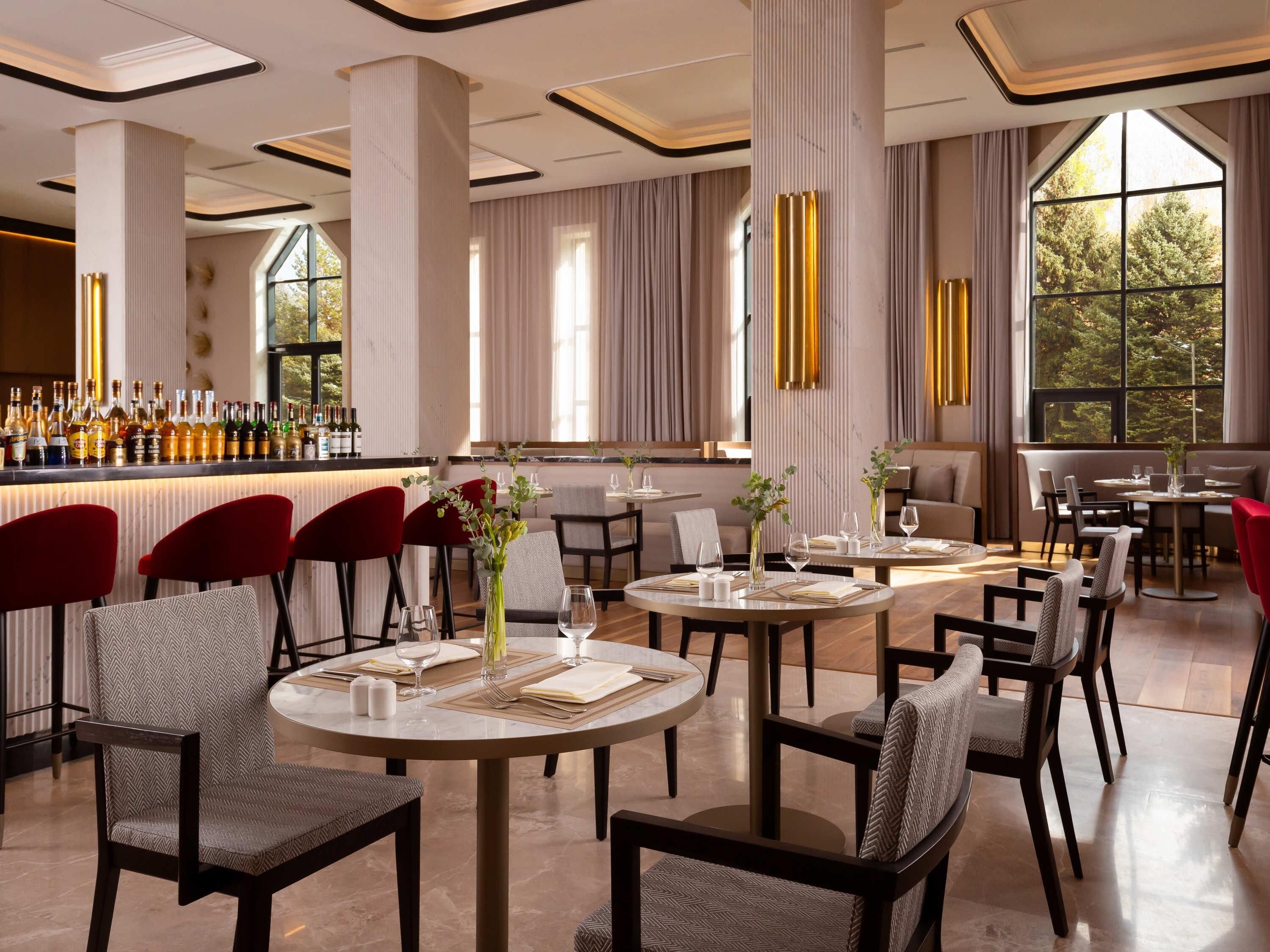 A modern restaurant with round tables, red chairs, and a bar area with bottles.