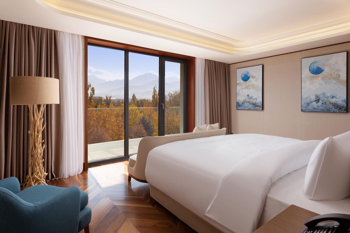 A hotel room with a large bed, a desk with a telephone, a chair, a lamp, and a view of trees and mountains through the window.