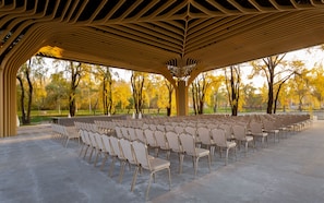 Un pavillon extérieur doté de rangées de chaises, d’un sol en béton et offrant une vue sur des arbres aux feuilles jaunes.