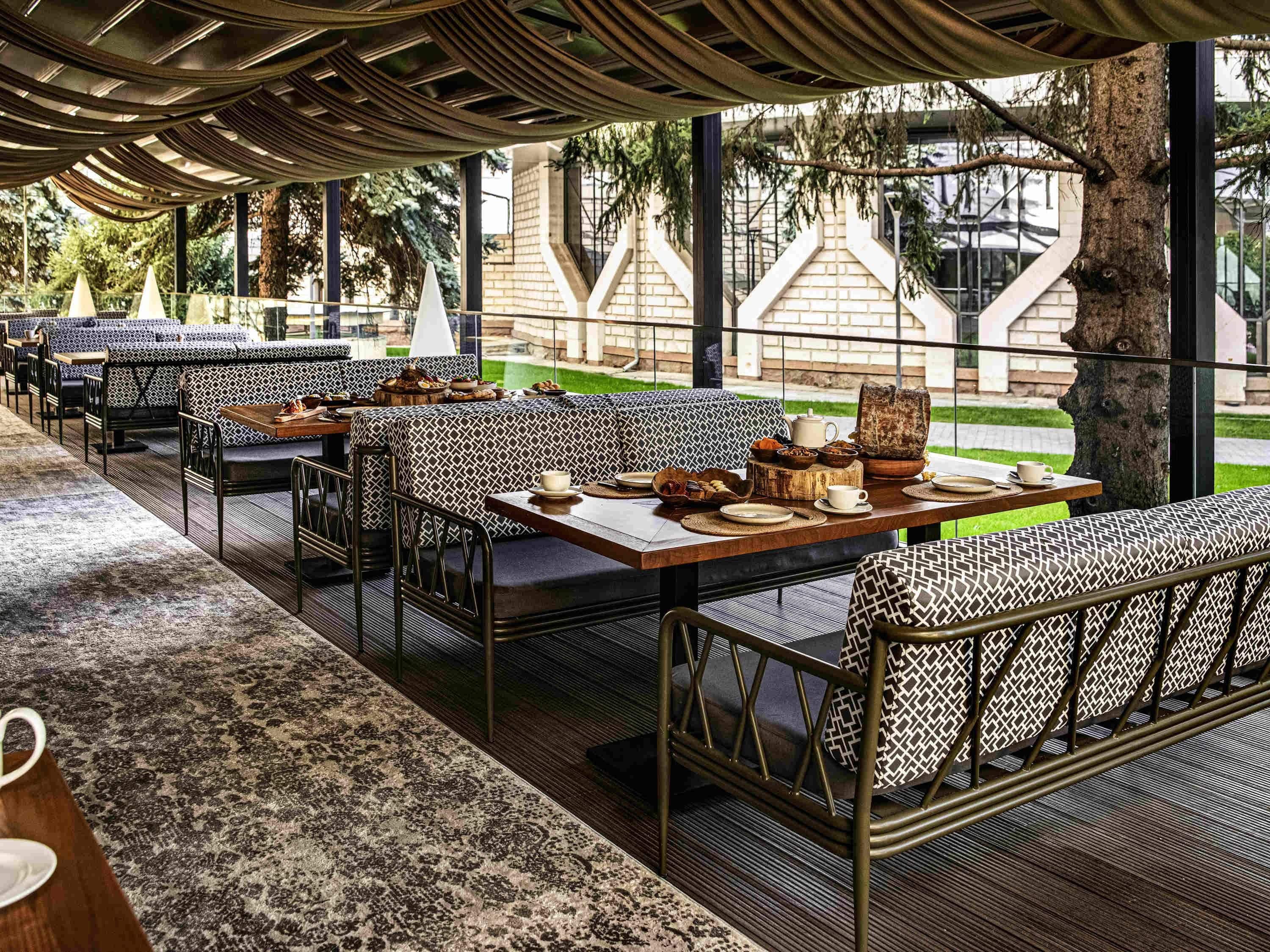 Un área de comedor al aire libre con asientos estampados, mesas dispuestas con vajilla y vistas a un edificio y árboles.