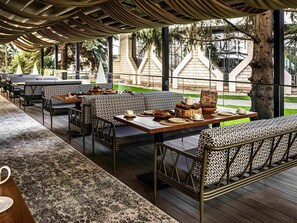 Een terras met patroonvormige zitplaatsen, tafels gedekt met servies, en uitzicht op een gebouw en bomen.