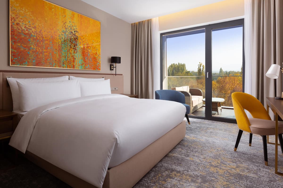 Une chambre d’hôtel avec un grand lit, un bureau, une chaise, une fenêtre donnant sur un beau paysage et un tableau au mur.