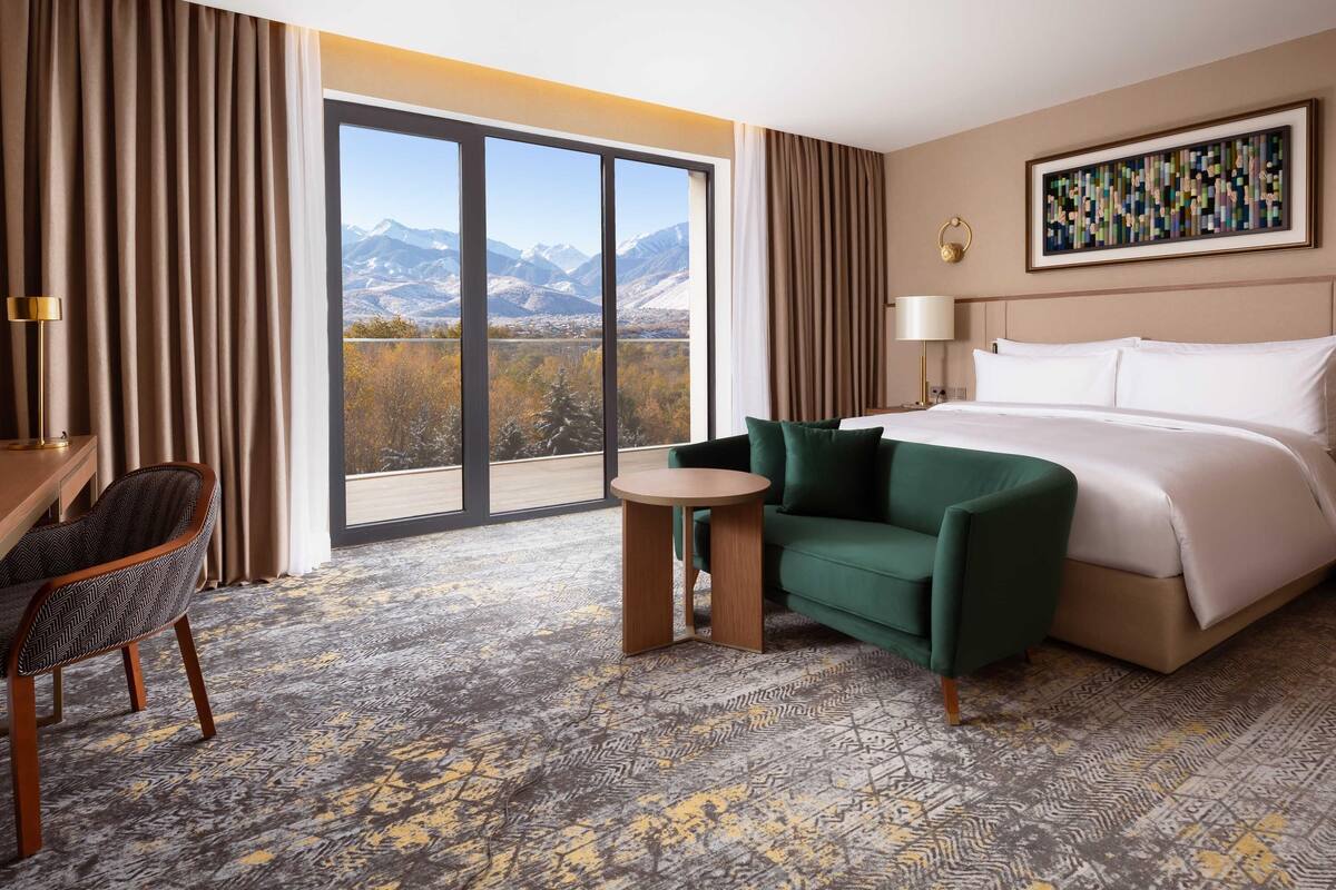 Une chambre d’hôtel avec un grand lit, un fauteuil vert, une petite table et une vue sur les montagnes grâce à de grandes fenêtres du sol au plafond.