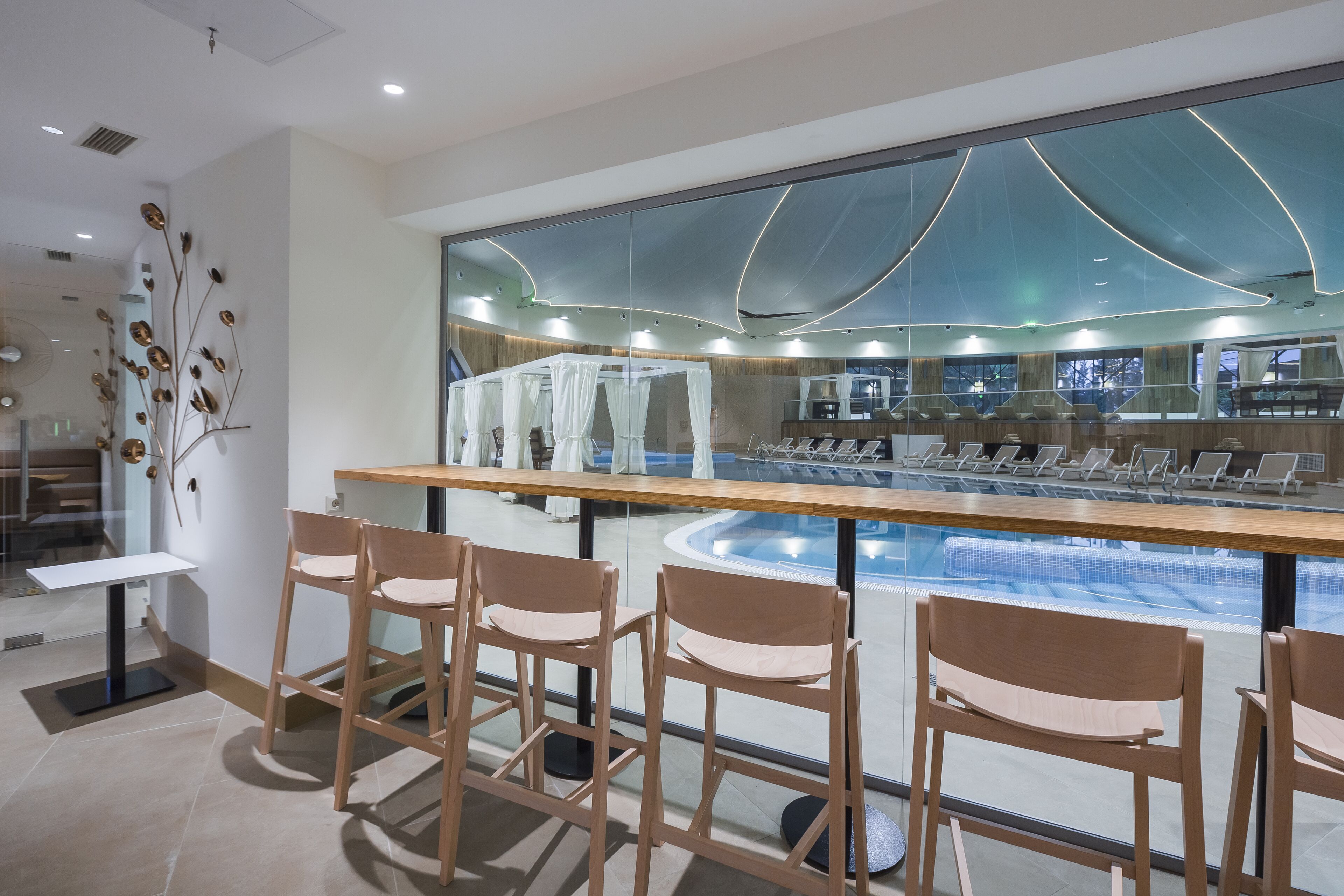 Eine moderne Bar mit Holzstühlen, Blick auf den Pool und einem großen Spiegel, der Pool und Deckenarchitektur reflektiert.