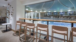 Un área de bar moderna con sillas de madera, vista a la piscina y un espejo grande que refleja la piscina y el diseño del techo.