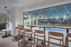 Et moderne barområde med træstole, udsigt til poolen og et stort spejl, der afspejler poolen og loftets design.