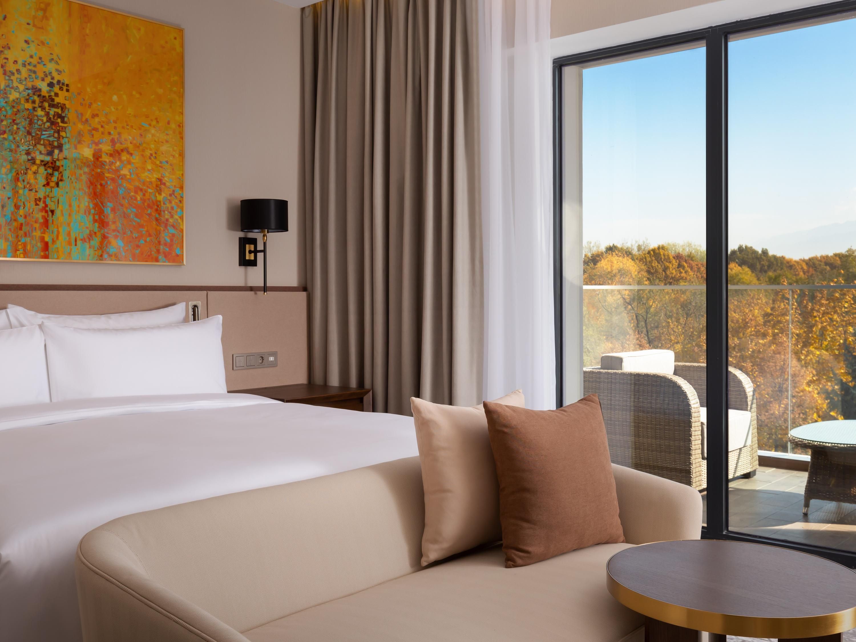 A hotel room with a bed, a sofa, a table, a lamp, and a view of the outdoors.