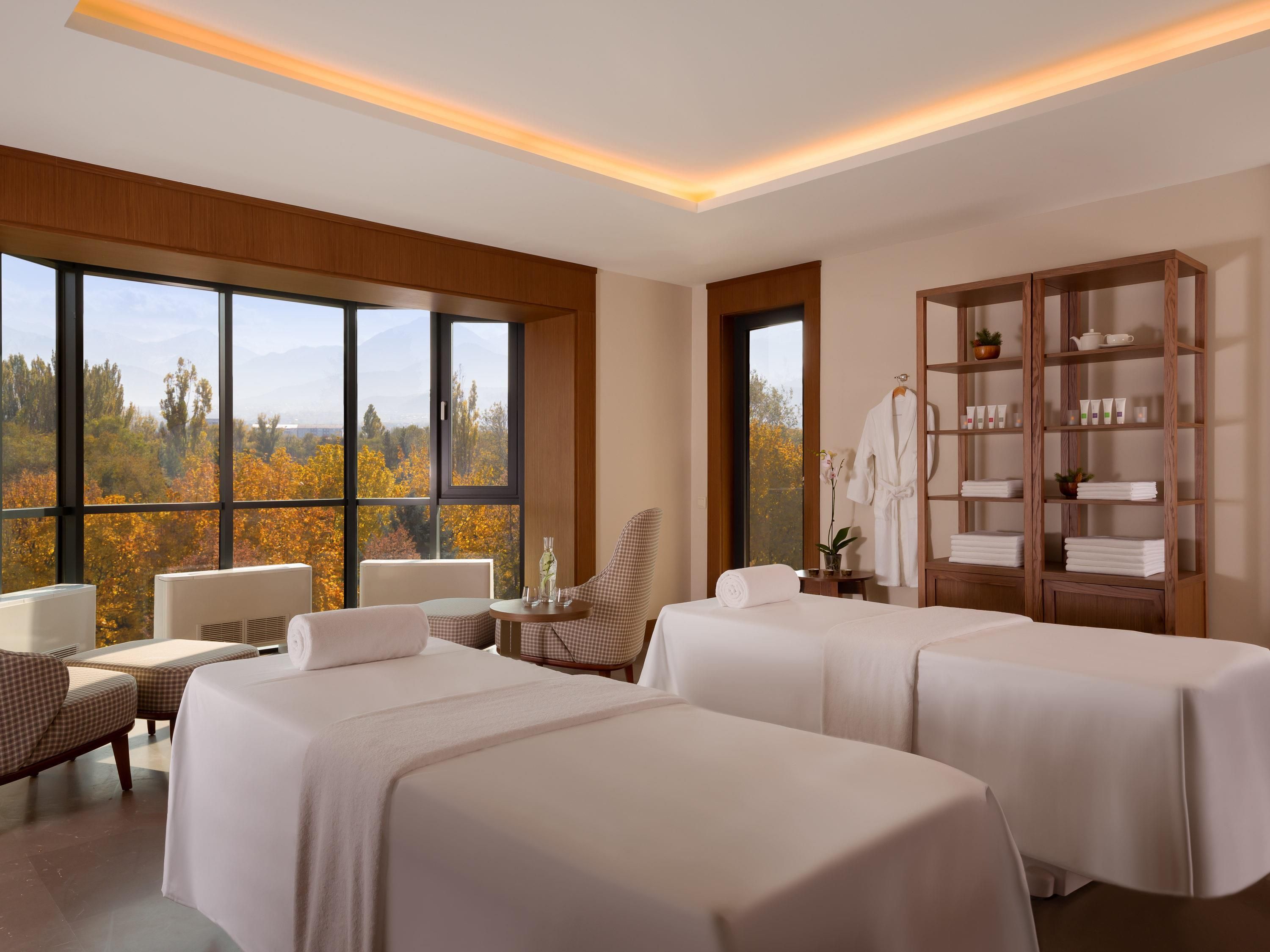 A hotel room with two beds, a seating area, a large window with a view of autumn trees, and a wooden cabinet with toiletries.