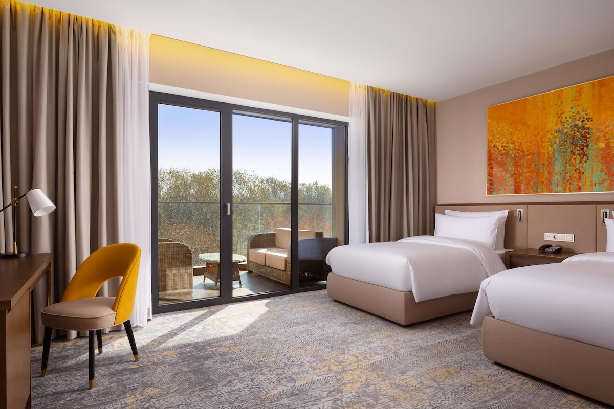 A hotel room with a balcony, two beds, a desk, a chair, and a large abstract painting.
