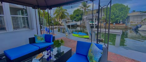 An outdoor seating area with blue cushioned sofas and a glass-top table, overlooking a water body with boats and a swing set.