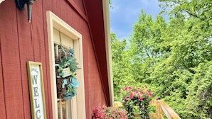 Een rood gebouw met een 'WELCOME' bord, een deur en een houten terras met een bankje en plantenpotten.