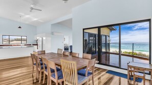 A modern kitchen with white cabinets, a dining area with a wooden table and chairs, and a large sliding glass door leading to a balcony with a view of the ocean. - Bermi Bliss - Dickinson Avenue 7 (Bermagui)