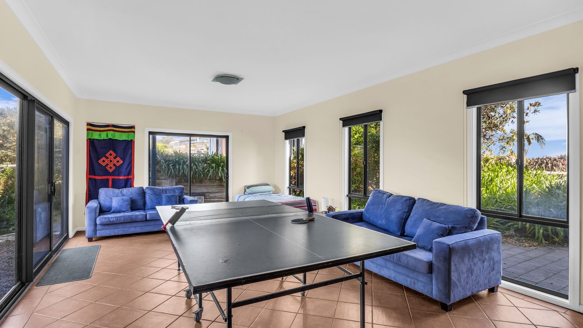 A room with a ping pong table, two blue sofas, and large windows offering a view of greenery.