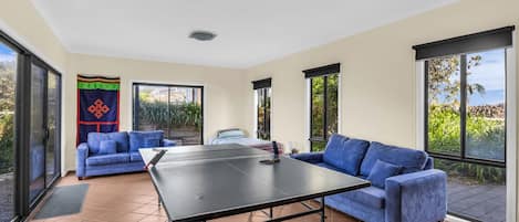 A room with a ping pong table, two blue sofas, and large windows offering a view of greenery.